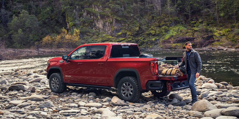A leather-jacketed guy is lifting camping gear out of the back of his red 2026 GMC Sierra 1500 as it is parked on the rocky shore beside a small smooth-flowing river in the mountains.