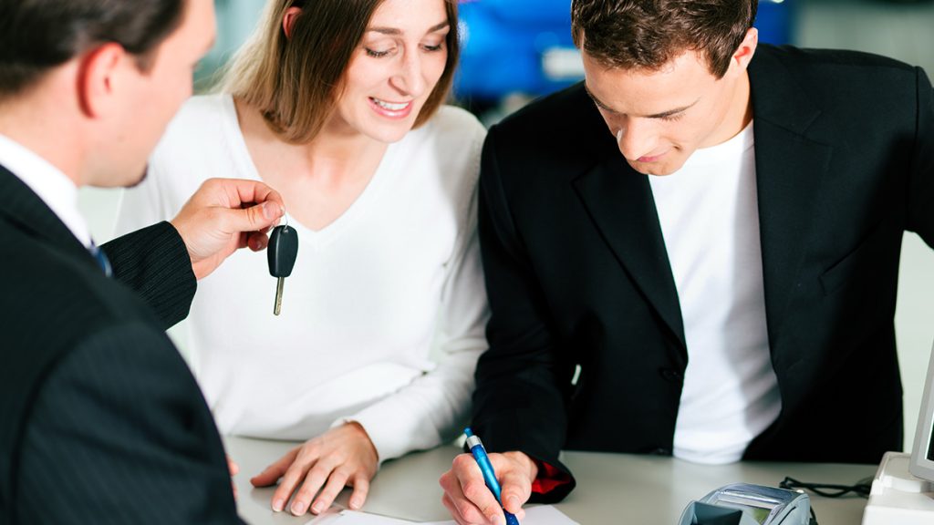 New Car Financing contract being signed by the husband of a smiling  couple as the sales representative hands them the key to their new vehicle.